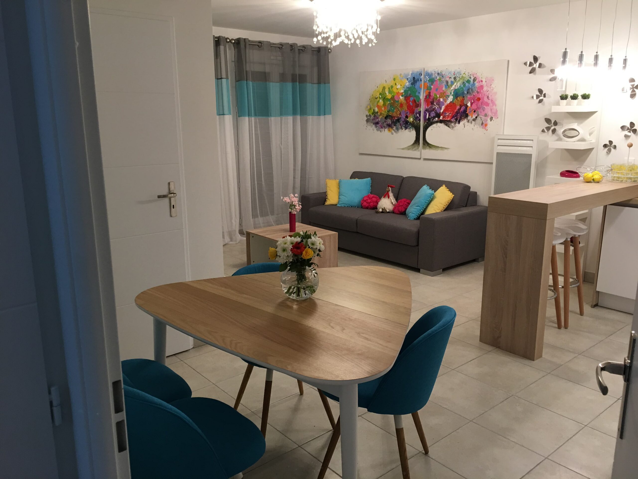 Salon moderne et lumineux avec canapé gris, touches turquoise et fuchsia, coin repas avec table en bois clair et coin cuisine avec bar.